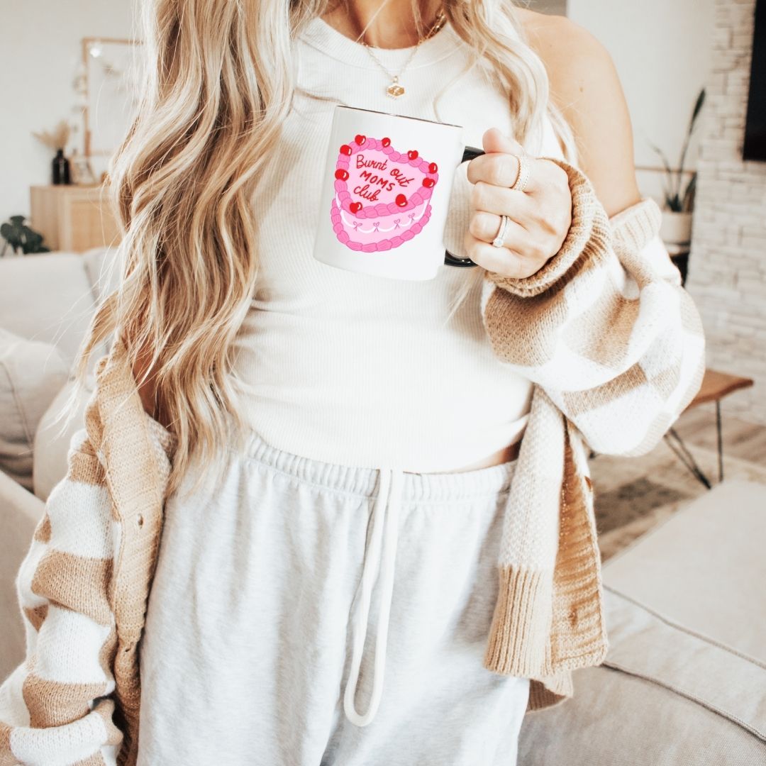 woman holding a white mug featuring a pink cake illustration with the text 'burnt out moms club'